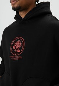 Zwarte hoodie gemaakt van een katoenmengsel. Voorzien van een afbeelding van een rode roos op de borst met tekst. Heeft een voorzak en geribbelde boorden.