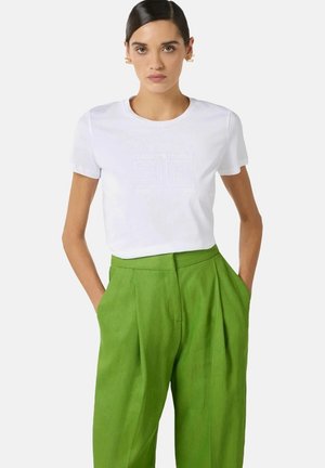 Donna con camicia bianca a maniche corte con logo discreto, pantaloni plissettati a vita alta verde brillante e orecchini geometrici dorati, mani nelle tasche.