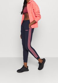 Felpa rosa sopra un reggiseno sportivo bordeaux, abbinata a pantaloni jogger navy con strisce laterali rosa e un logo. Scarpe da ginnastica nere completano l'outfit.