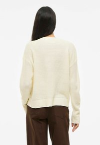 Pull en tricot crème avec une coupe ample, des épaules tombantes et une texture côtelée. Présente un ourlet court et des manches longues. Associé à un pantalon marron.