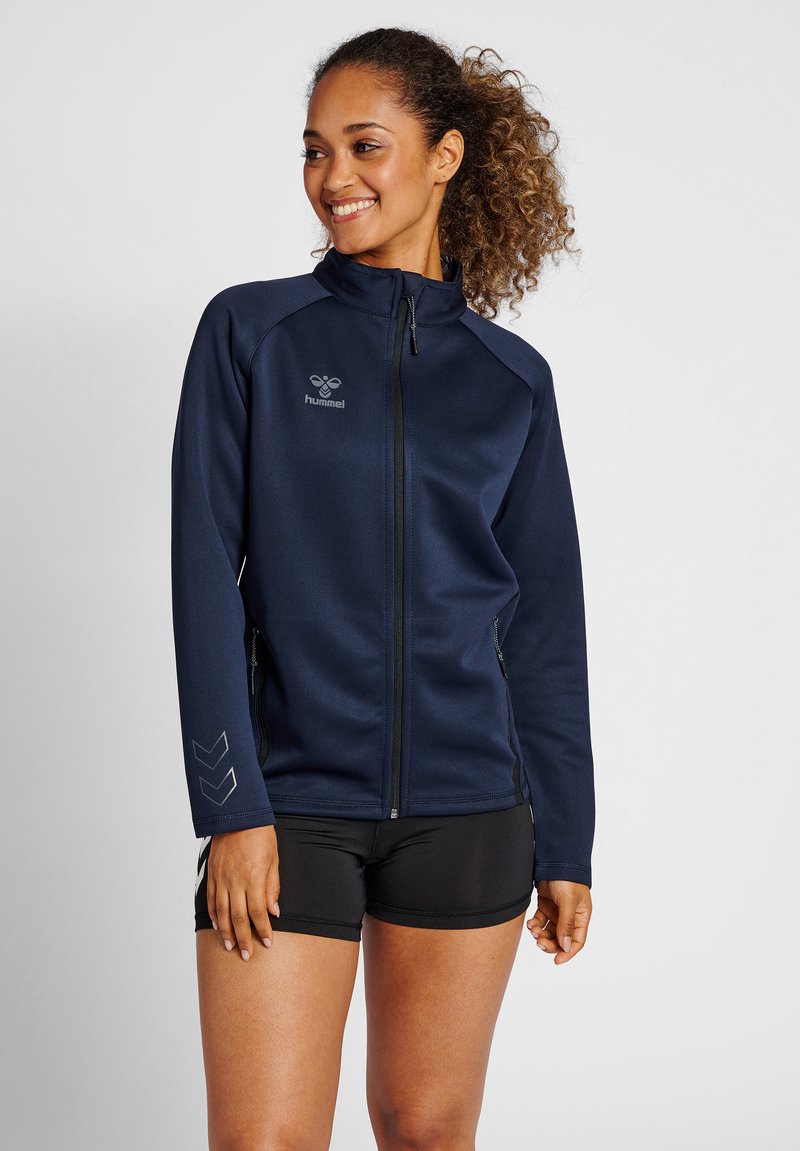 Frau mit lockigem Haar, die eine marineblaue Sportjacke mit Reißverschluss und schwarze Sportshorts trägt, steht und lächelt, leicht nach links schauend.