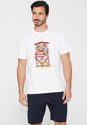 Mann trägt weißes T-Shirt mit Cartoon-Bär im Rettungsturm und marineblaue Shorts, steht vor schlicht weißem Hintergrund.