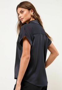 Schwarze Satinbluse mit kurzen aufgerollten Ärmeln, einem Kragen und einem leicht lockeren Schnitt. Glatte Textur mit einem klaren, minimalistischen Design.