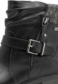 Black faux leather ankle boot with a decorative strap and silver buckle, featuring a textured fabric panel and side zipper.