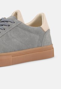 Γκρι σουέντ αθλητικό παπούτσι με μπεζ ταμπά στην φτέρνα, σκούρα γκρι κορδόνια και σόλα από λάστιχο τύπου καουτσούκ σε λευκό φόντο.