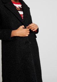 Manteau en laine noire texturée avec un col cranté, une fermeture à un bouton et deux poches latérales. Superposé à un pull rayé rouge.