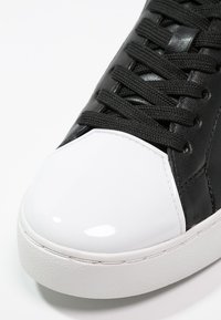 Gros plan d’une basket noire avec un embout blanc brillant et des lacets noirs, montrant une texture détaillée et des coutures sur fond blanc.