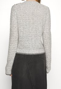 Hellgrauer Strickpullover mit geripptem Saum und Ärmeln, strukturiertem Muster und dekorativen Schulterknöpfen. Kombiniert mit einem dunkelgrauen Rock.