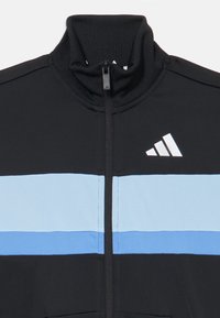 Črna športna jakna s stoječim ovratnikom. Ima svetlo modro vodoravno črto in belo logo na levi strani. Spredaj z zadrgo.