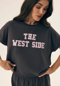 Dark gray cotton t-shirt with wide sleeves, featuring "THE WEST SIDE" in bold white and pink letters across the front. Soft texture.
