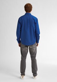 Modrá dlouhá košile s knoflíkovým zapínáním, která má špičatý límec a manžety. Kombinováno se světle šedými džíny a bílými teniskami.