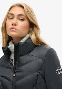 Chaqueta de plumas acolchada negra con collar alto, borde de forro polar gris y logo en la manga. Patrón de chevrón notable en la tela.