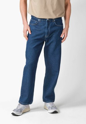 Persoon die blauw rechte jeans, een beige shirt en grijs-witte sneakers draagt, staand tegen een effen lichte achtergrond.