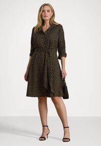 Rochie neagră cu un model circular auriu, cu nasturi pe față și talie legată. Are mâneci trei sferturi și o fustă evazată. Pantofi cu toc negri, cu bretele.