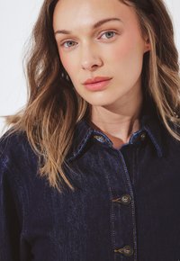 Vrouw met lichtbruin haar die een donkerblauwe denimjas met knopen draagt, recht in de camera kijkend tegen een witte achtergrond.