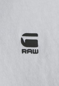 Vitt bomullstyg med en svart logotyp som visar stiliserade bokstäver "G" och "RAW". Designen har en slät yta och en minimalistisk estetik.