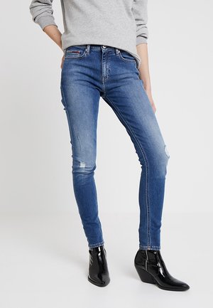 Person iført blå slim-fit jeans med let slid, grå langærmet top og sorte skinnende ankelstøvler med kilehæl, stående på hvidt gulv.