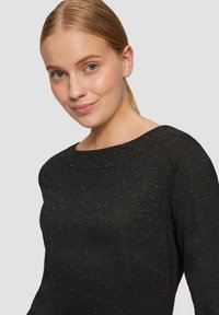 Zwarte gebreide top met een ronde halslijn, voorzien van een textuurpatroon. De stof heeft een lichte glans en subtiele onregelmatigheden, wat de visuele interesse vergroot.