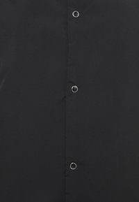 Czarna koszula o gładkiej fakturze, z guzikiem na przodzie i połyskującymi metalowymi guzikami. Tkanina wydaje się trwała i przewiewna.