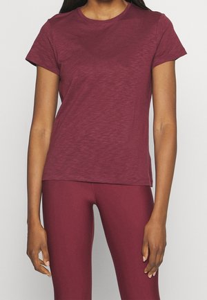 Kvinde iført en kortærmet vinrød t-shirt og matchende vinrøde leggings, stående med armene afslappet ned langs siderne.