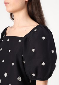Blusa nera con scollatura quadrata, maniche a sbuffo e ricami floreali bianchi. Tessuto morbido e dettagli arricciati alle aperture delle maniche.
