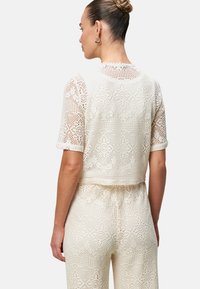 Top corto in pizzo bianco con maniche corte, caratterizzato da intricati motivi all'uncinetto e bordi a scallop sulla scollatura. Vista posteriore visualizzata.
