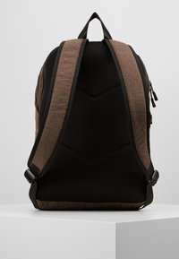 Sac à dos marron et noir avec un dos en maille texturée, des bretelles rembourrées et un devant en tissu lisse. Comprend des fermetures éclair et une solide poignée supérieure.