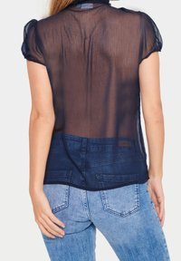 Donna che indossa una camicetta trasparente blu navy a maniche corte sopra una maglietta scura, abbinata a jeans di jeans azzurri, vista da dietro.