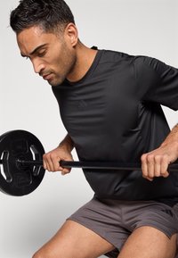 Μαύρο αθλητικό μπλουζάκι με κοντά μανίκια, με διακριτικό λογότυπο. Γκρι σορτς από δίχτυ, κρατώντας μια μαύρη δίσκο μπάρα κατά την εκτέλεση μιας άσκησης.