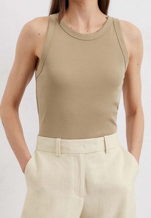 Donna con un top beige senza maniche infilato in pantaloni crema a vita alta con le mani in tasca, su uno sfondo chiaro e semplice.