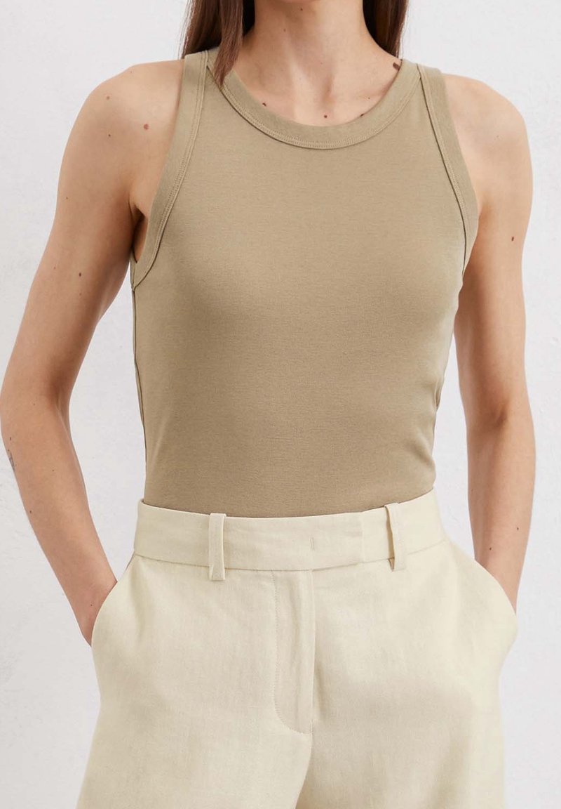 Donna con un top beige senza maniche infilato in pantaloni crema a vita alta con le mani in tasca, su uno sfondo chiaro e semplice.