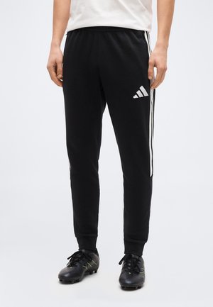 Osoba ubrana w czarne spodnie dresowe Adidas z białymi paskami i czarne korki, stojąca na jednolitym tle.