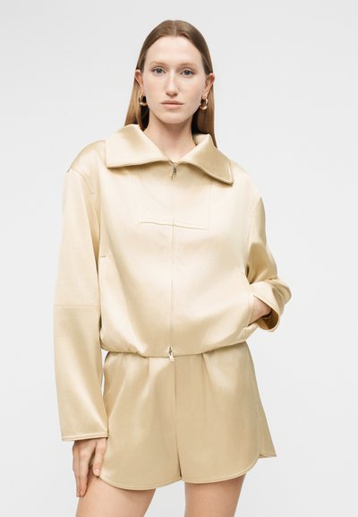 Γυναίκα με ίσια ανοιχτό καστανά μαλλιά φοράει μπεζ φαρδιά ζακέτα με φερμουάρ και ταιριαστό σορτς, στέκεται με το ένα χέρι στην τσέπη.