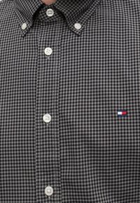 Chemise à petits carreaux noirs et gris avec des boutons blancs et un petit logo rectangulaire rouge, blanc et bleu sur la poitrine.