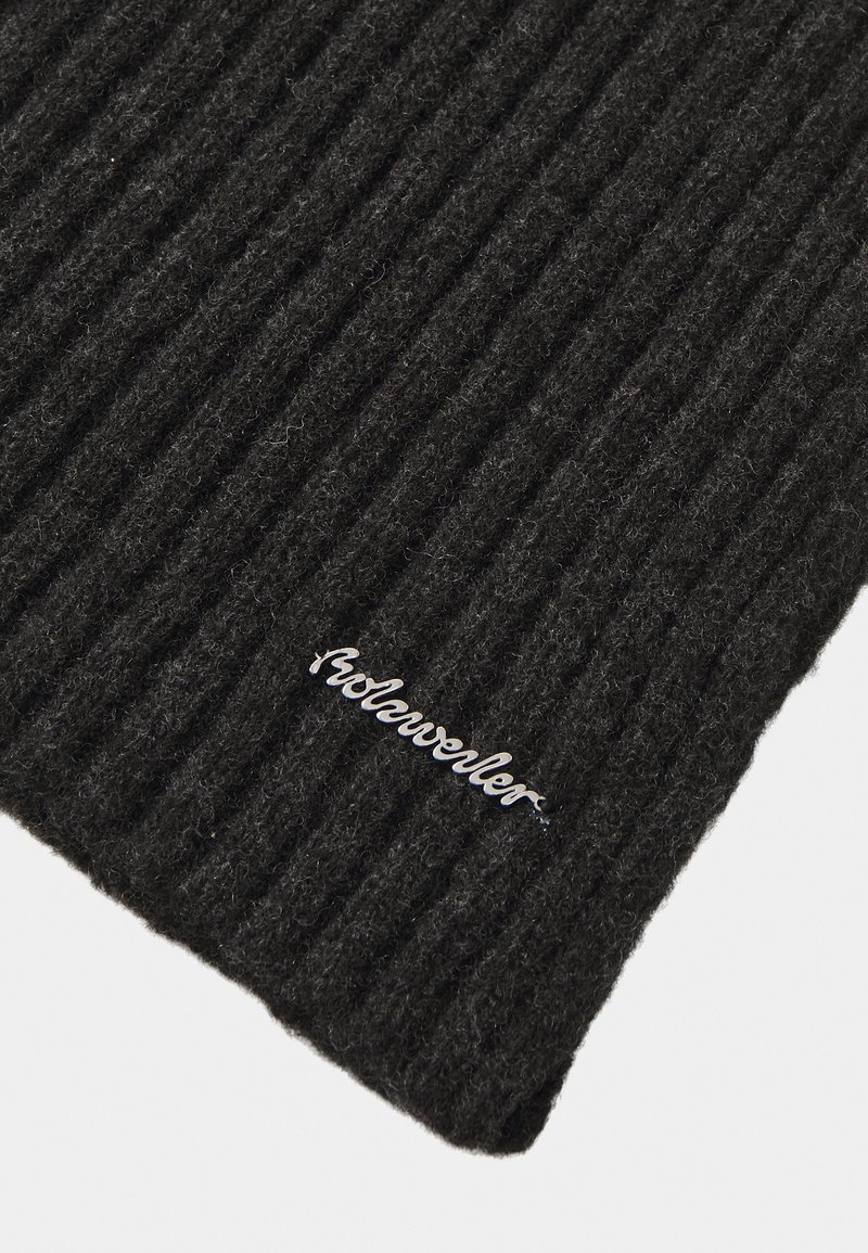 Béret en tricot noir à côtes, fabriqué en laine ; présente une étiquette logo argentée cousue sur le bord. La texture est douce et chaude.