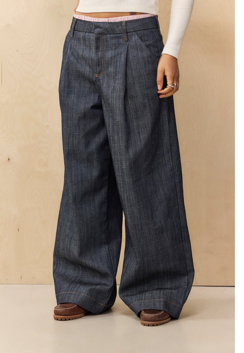 Pantalones de mezclilla de pierna ancha en azul oscuro, con pliegues, una tela texturizada y costuras en contraste. Combinados con calzado marrón.