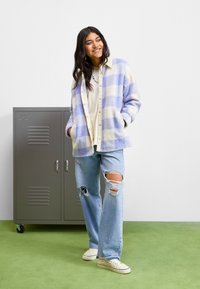 Vloeiende geruite jas in lichtpaars en roomkleur, met knopen aan de voorkant, patchzakken en een ontspannen pasvorm, gedragen over een roomkleurig topje en versleten spijkerbroek.