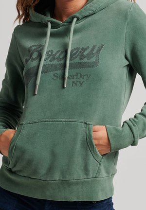 Osoba ubrana w zieloną bluzę z kapturem SuperDry z trokami i przednią kieszenią typu kangur, ręce schowane w kieszeni.