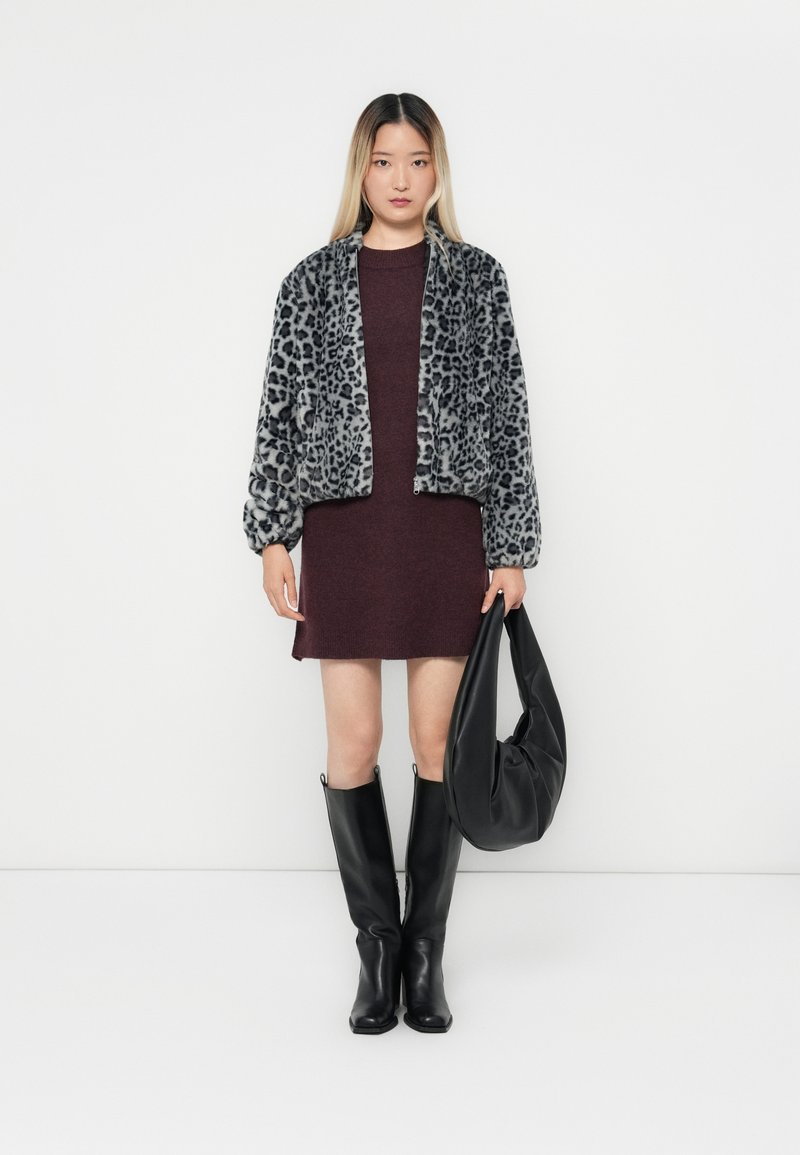 Grau gefleckte Kunstfell-Bomberjacke über einem kastanienbraunen Kleid, kombiniert mit knielangen schwarzen Stiefeln und einer lässigen schwarzen Handtasche.