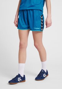 Osoba v modrých sportovních šortkách a odpovídajících teniskách s bílými ponožkami, stojící na jednolitém, světlejším pozadí.