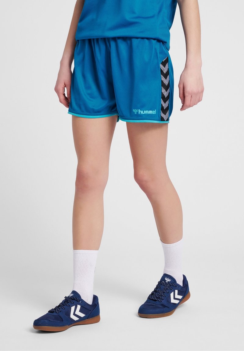 Henkilö, jolla on yllään siniset urheilushortsit ja niihin sopivat tennarit valkoisten sukkien kanssa, seisoo yksinkertaisella, vaalealla taustalla.