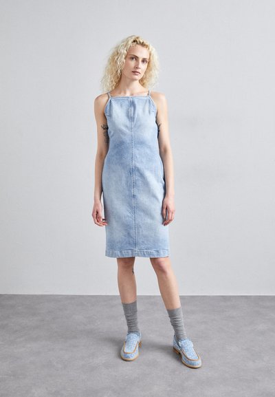 Denim haljina svijetloplave boje, bez rukava, ravnog kroja s prednjim šavom. Uparena s sivim čarapama i svijetloplavim mokasinkama.
