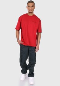 Rød bomulds T-shirt med korte ærmer og et lille sort logo, parret med mørke cargo bukser med sidelommer og hvide sneakers.