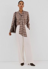 Geruite blouse in bruintinten met een geknoopte halslijn; gecombineerd met hooggetailleerde crèmekleurige broek en puntige bruine hakken. Bruine schoudertas.