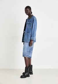 Casaco de ganga com colarinho castanho, duas bolsos frontais, combinado com uma saia de ganga até ao joelho e botas pretas até ao meio da panturrilha com fivelas.