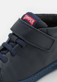 Tmavě modrá kožená vysoká bota s suchým zipem, strukturovaným povrchem a modrou podrážkou. Logo Camper v červené barvě na kotníku.