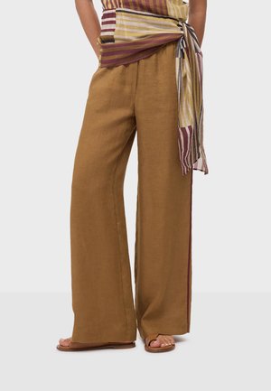 Pantalon ample en lin marron porté avec une ceinture à nouer rayée et des sandales plates ouvertes, visible de la taille aux pieds sur un fond clair.