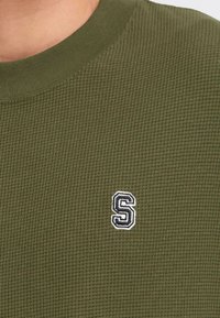 Olive green, textured knit sweater with a round neckline and small black-and-white embroidered 'S' logo on the left chest.