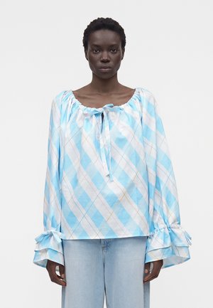 Kvinde iført en løs, langærmet blå og hvid ternet bluse med rynkede manchetter og lyseblå jeans, der vender front mod en hvid baggrund.
