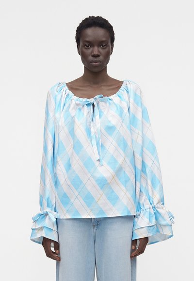 Femme portant un chemisier ample à manches longues à carreaux bleus et blancs avec des poignets à volants et un jean bleu clair, faisant face à l'avant sur fond blanc.